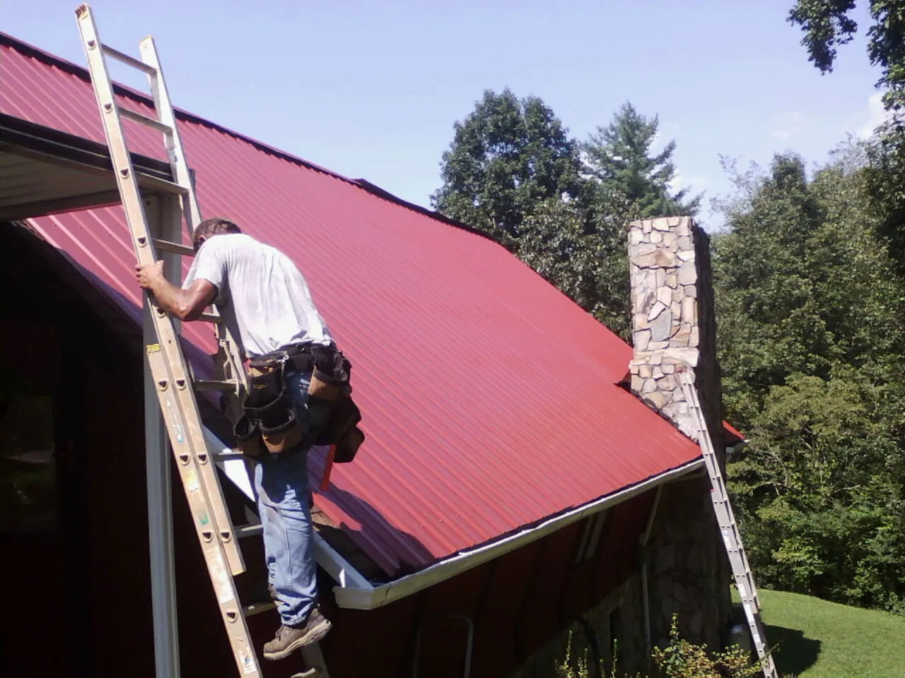 Roofer on a ladder beside a red metal roof and stone chimney — Soffit & Fascia Repair by Roofing Brothers in Clarkia